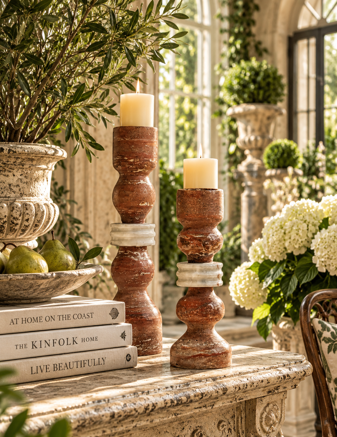 Two rustic terracotta candlestick holders with white stone accents, each holding a white pillar candle, styled on a stone mantel in a sunlit interior with greenery and flowers.