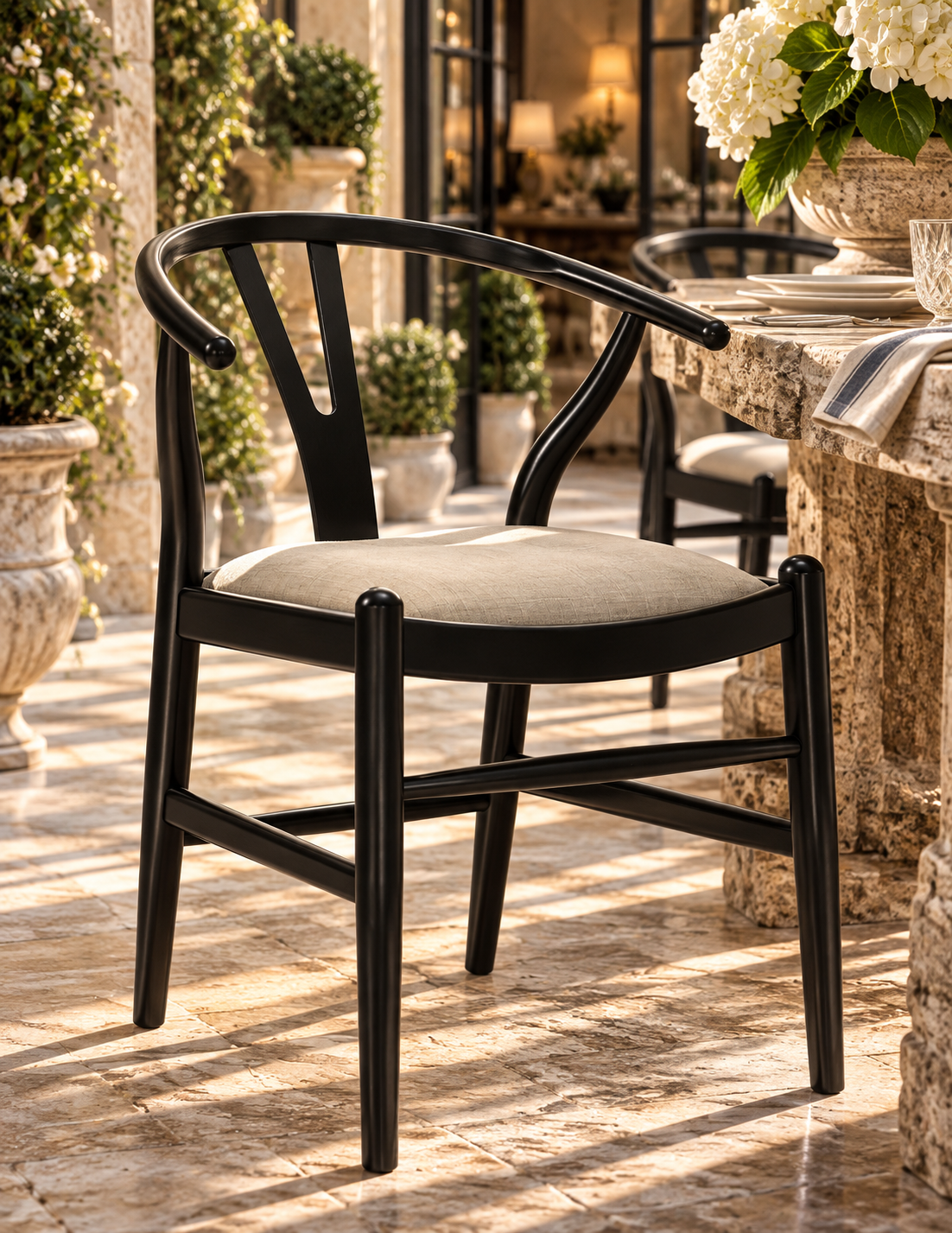 A black wood dining chair with a curved backrest and a light beige upholstered seat, shown on a sunlit stone patio beside a table with flowers.