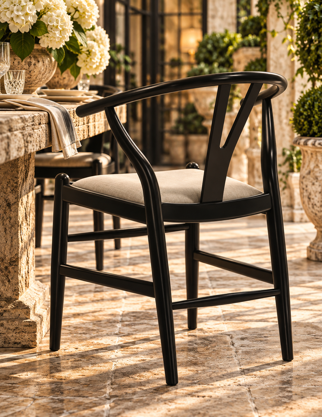 A black wooden armchair with a light beige cushioned seat, positioned on a stone tiled patio next to a stone table with white flowers.