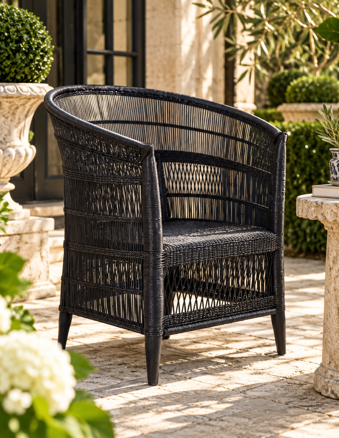 A black woven wicker armchair with a curved backrest, shown on a stone patio in a garden setting.