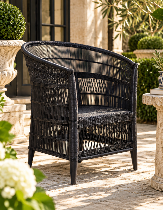 A black woven wicker armchair with a curved backrest, shown on a stone patio in a garden setting.