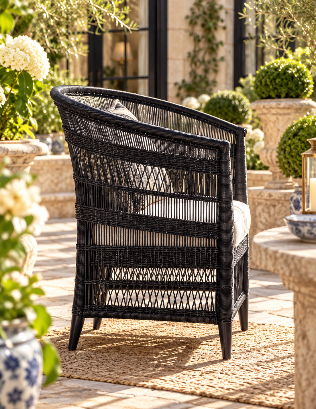A black woven outdoor armchair with a curved backrest and cream cushions, placed on a woven rug in a sunlit garden patio setting.