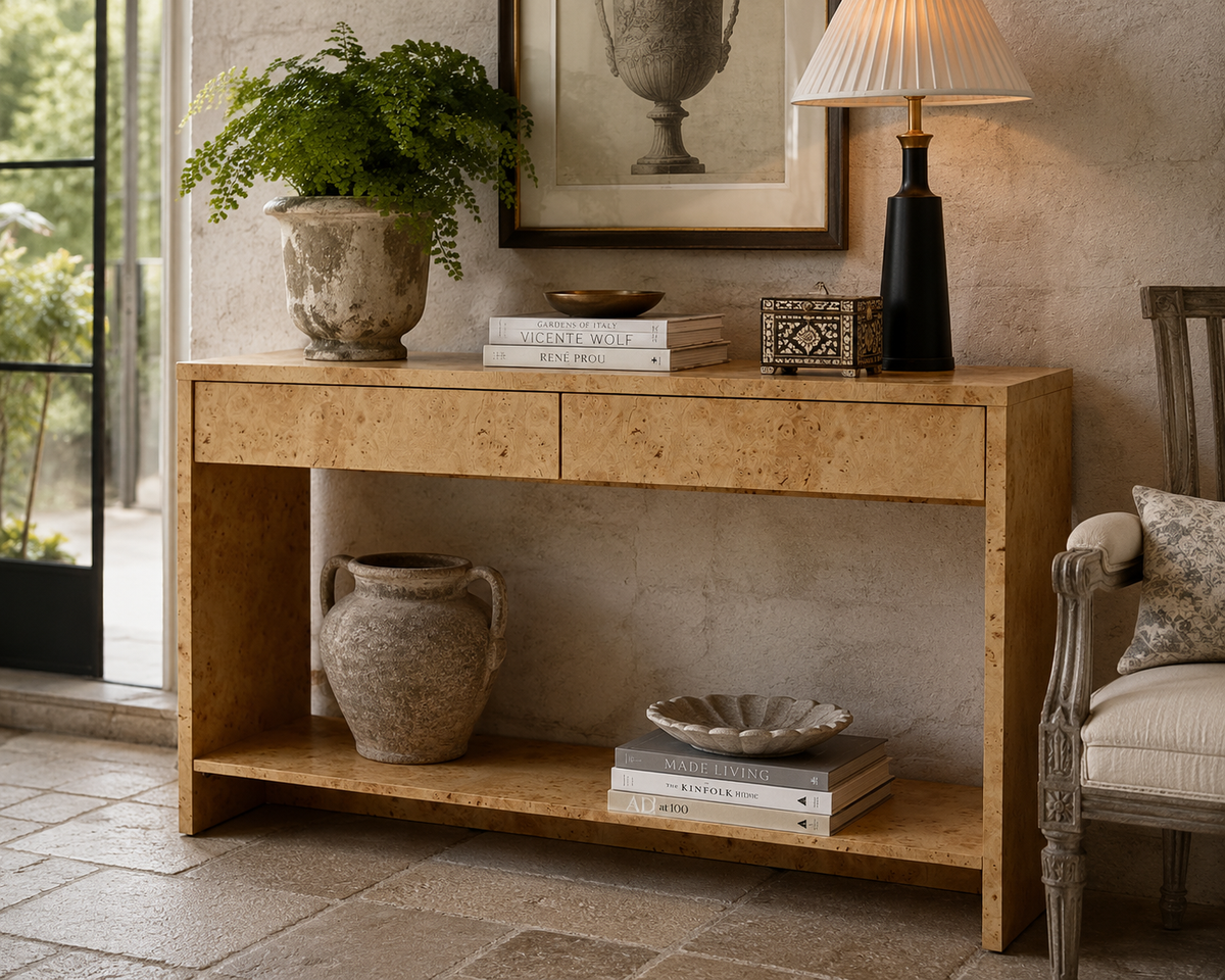 A light natural wood console table styled in a rustic room setting, featuring a top drawer, open lower shelf, and decorative items including a potted fern, books, a ceramic vase, and a table lamp.