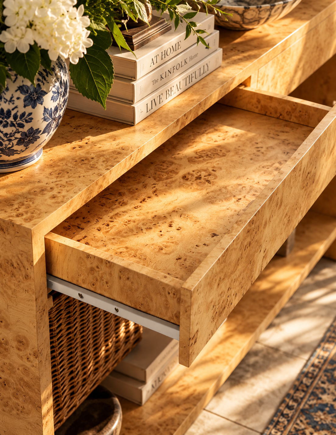 A close-up of a light natural wood drawer pulled open on a matching wooden console table, with books, a blue-and-white floral vase, and sunlight casting warm shadows across the drawer interior.