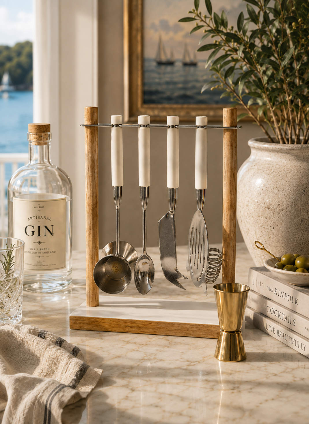 A wooden bar tool holder with five white-handled utensils, displayed on a marble countertop in a styled kitchen scene next to a gin bottle, books, olives, and a vase.