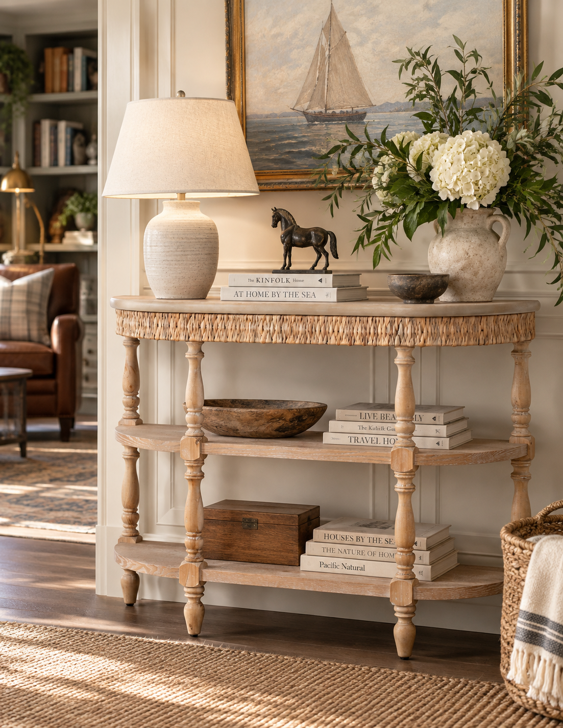 A rustic console table styled in a cozy living room setting, featuring a woven top, turned wood legs, and open shelves. The table is decorated with a textured ceramic table lamp, a small black horse figurine, books, a wooden bowl, and a large ceramic vase filled with white flowers.