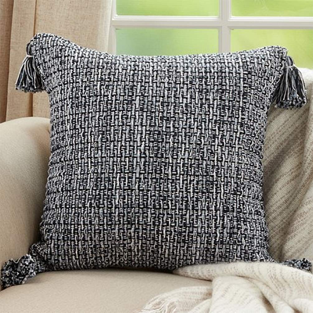 A square woven throw pillow in a black and white textured pattern, styled on a beige armchair with a cream knit blanket and tassels on the corners, set in front of a window.