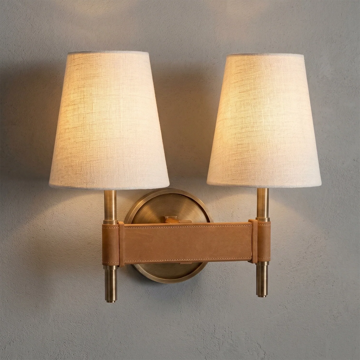 Wall-mounted lamp with two beige lampshades and a brown leather strap on a gray wall.