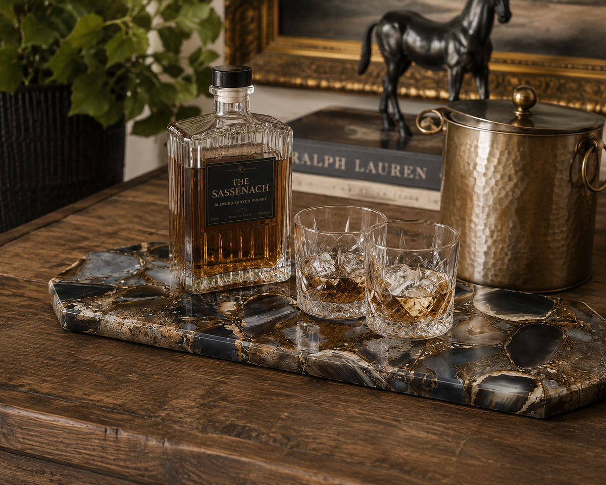 A whiskey bottle, two rocks glasses, and a hammered metal bucket on a decorative marble serving tray, arranged on a rustic wooden table in a styled interior setting.