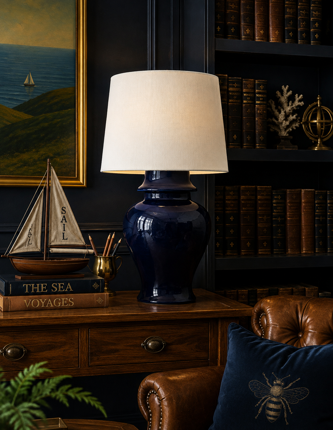 A navy blue ceramic table lamp with a large cream fabric drum shade, styled on a wooden console table in a dark, elegant room with a leather armchair, bookshelves, and a nautical pillow.