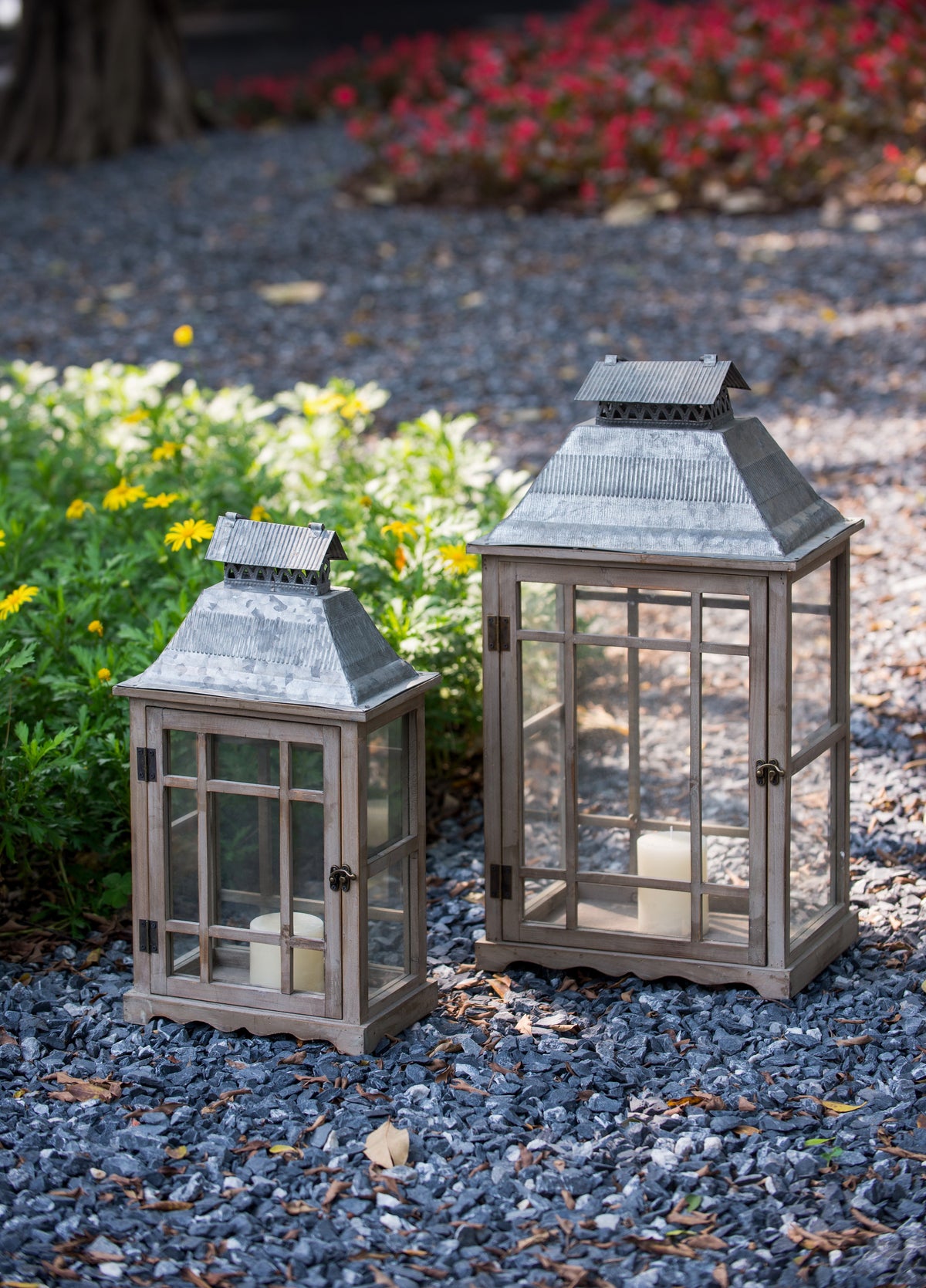 Two rustic metal lanterns with glass panels, one small and one large, placed on a gravel path in a garden setting.