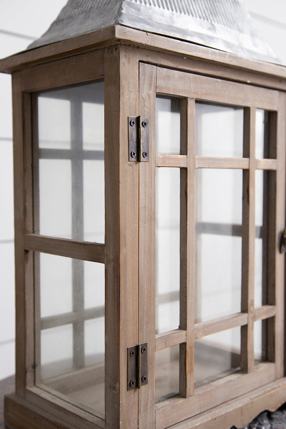 A rustic wooden lantern with a weathered brown finish, featuring glass window panes and dark metal hinges, shown in a close-up shot.