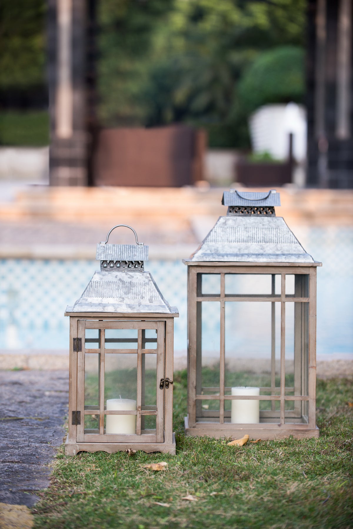 Two rustic metal lanterns with glass panels, one small and one large, placed on grass outdoors near a pool and blurred patio background.