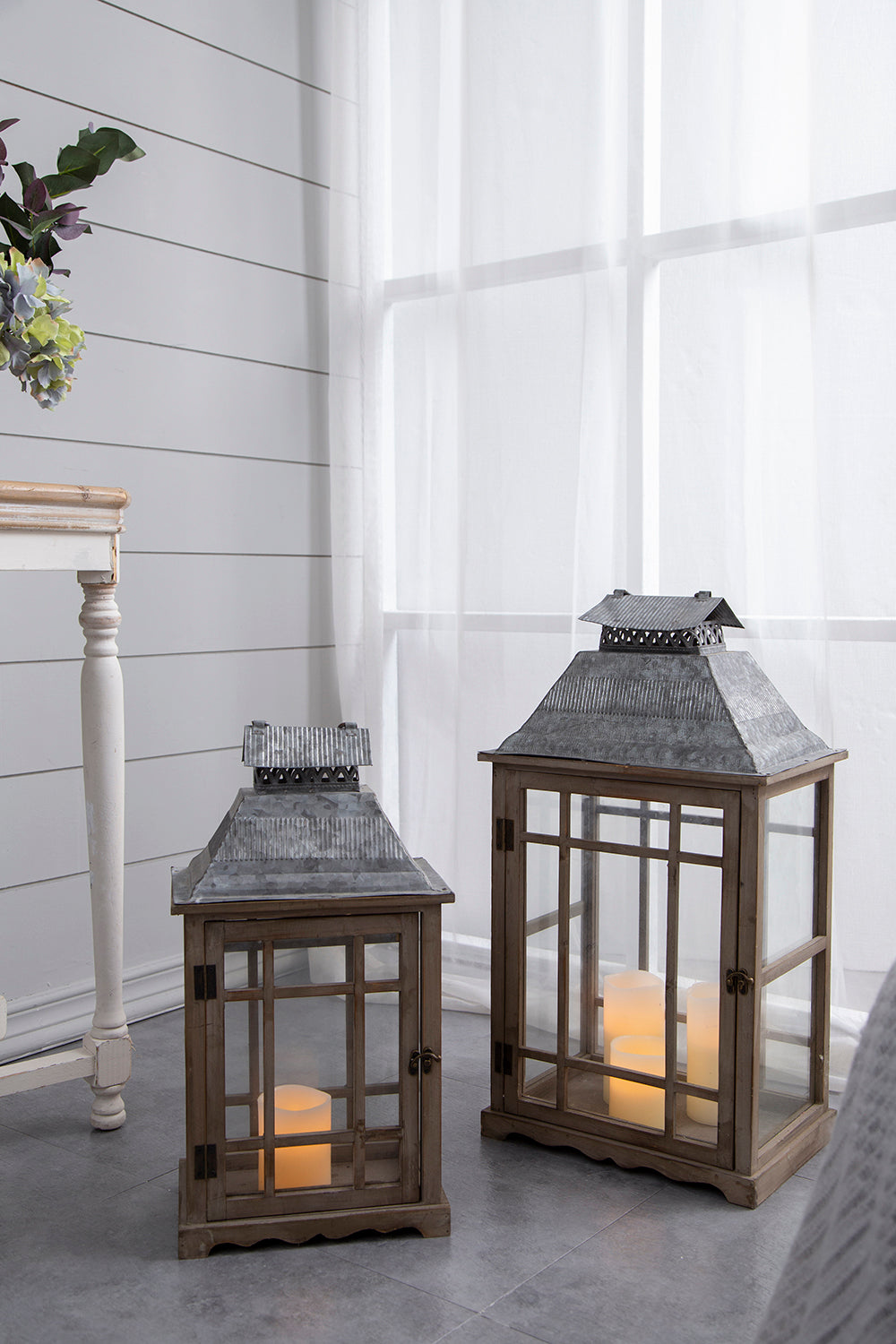Two rustic wooden lanterns with galvanized metal roofs, one small and one large, both lit with warm yellow candles, set on a gray floor against a white shiplap wall and sheer window curtains.