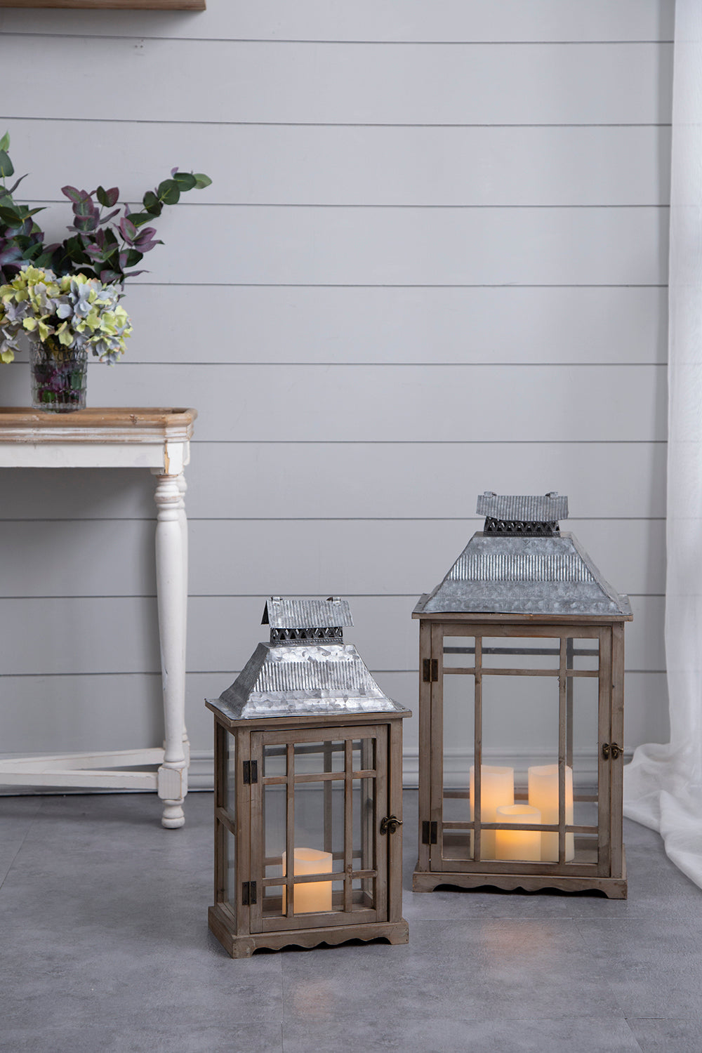 Two rustic wooden lanterns with galvanized metal roofs, one small and one large, both lit with warm candlelight, set against a light gray shiplap wall.