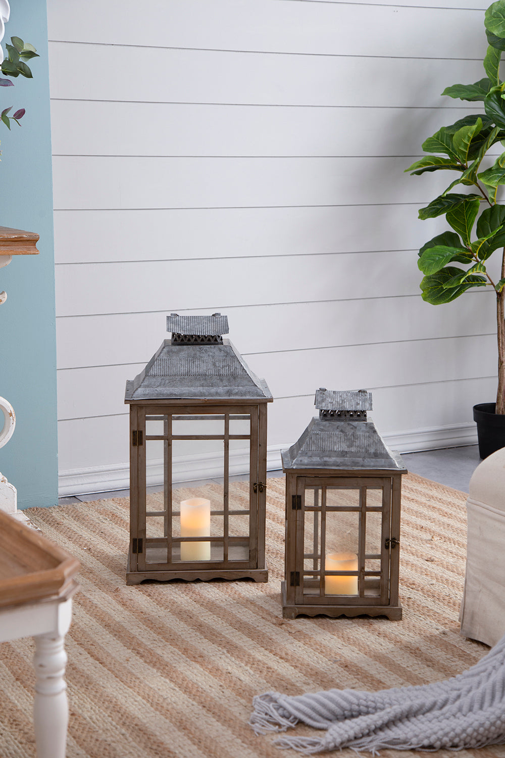 Two rustic wooden lanterns with gray metal roofs, one large and one small, each containing a lit candle, placed on a striped jute rug in a styled room setting.
