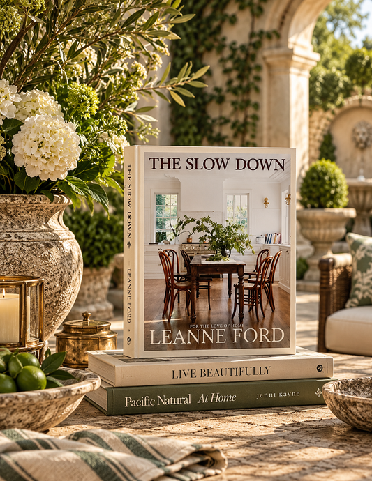 A book titled "The Slow Down" by Leanne Ford stands upright on top of two other books on a stone patio. The scene is styled with large floral arrangements in a rustic ceramic vase, candles, bowls, and outdoor seating in the background.
