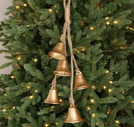 Four small gold bell Christmas tree ornaments hanging from natural jute rope on a green Christmas tree with warm white lights.