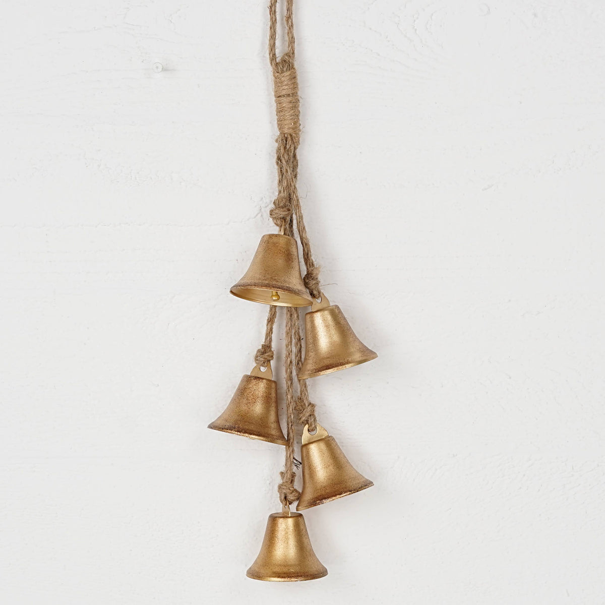 A hanging decorative garland made of five small metallic bells in a warm gold finish, suspended from a natural jute rope against a white textured wall.