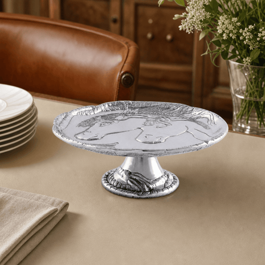 A silver pedestal cake stand with embossed horse designs on the top and base, displayed on a beige tablecloth in a dining setting with a brown leather chair and wooden cabinet in the background.