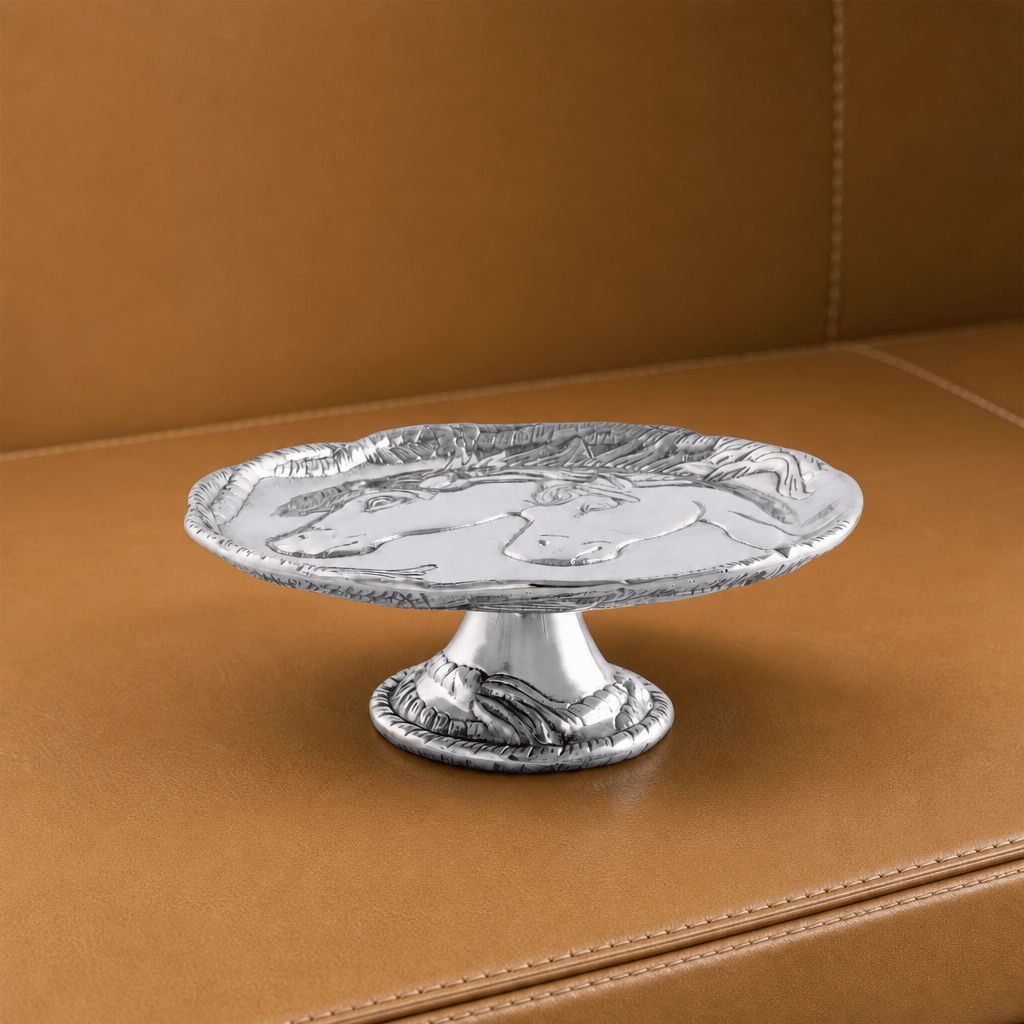 A silver pedestal stand with an embossed horse design on the round tray, displayed on a brown leather surface.