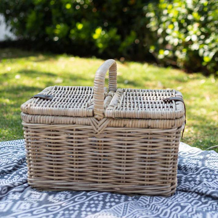 A rectangular woven wicker picnic basket with a top handle, placed on a blue and white patterned blanket outdoors on grass with a blurred green garden background.