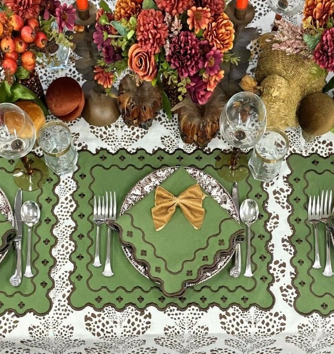 An overhead view of a decorated table setting featuring a green lace table runner with scalloped edges, white lace placemats, silver cutlery, clear wine glasses, and autumn floral arrangements with pumpkins and burlap decor.