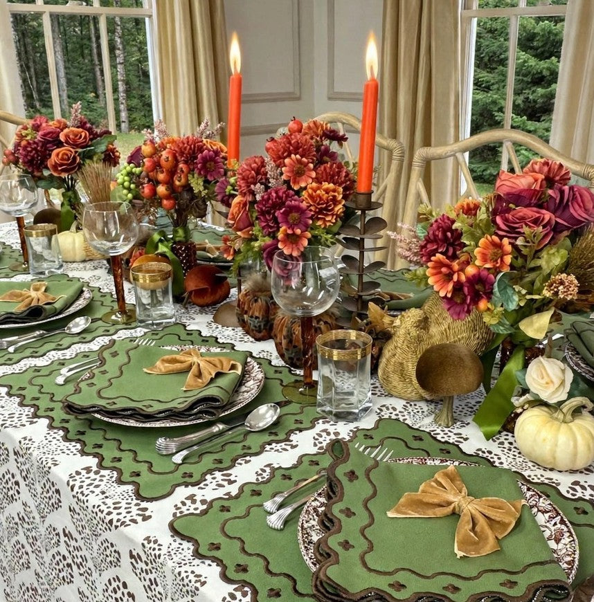 An autumn-themed table setting featuring olive green embroidered place mats with scalloped edges, matching green napkins tied with tan bows, white lace tablecloths, and vibrant fall floral centerpieces with orange and red candles.