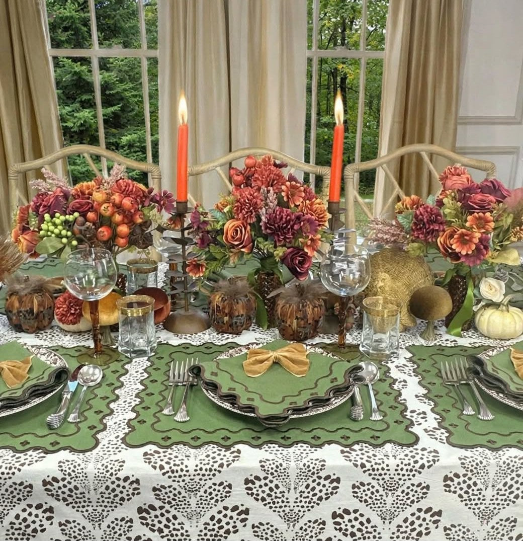 An autumn-themed table setting with olive green scalloped placemats, white lace tablecloths, layered plates, silverware, orange taper candles, and floral centerpieces in fall colors, set in a dining room with window views.